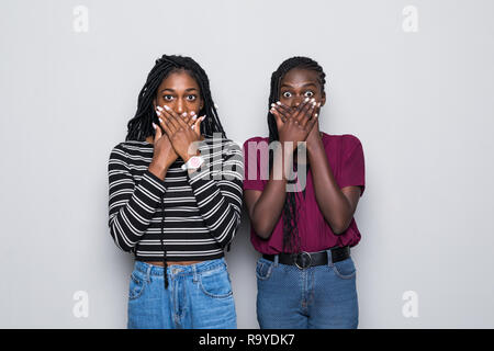 Waist up shot of shocked two african women cover mouthes with both hands, try be speechless in stressful situation isolated over gray background. Stock Photo
