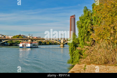 SEVILLE SPAIN THE SEVILLA OR CAJASOL TOWER WITH TOURIST BOAT ON THE GUADALQUIVIR RIVER PASSING UNDER THE TRIANA METAL ARCH BRIDGE Stock Photo