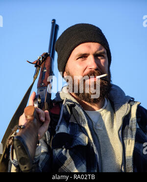 Hunter with rifle gun close up. Guy bearded hunter spend leisure hunting and smoking. Hunting masculine hobby concept. Man brutal bearded guy gamekeeper blue sky background. Brutality and masculinity. Stock Photo