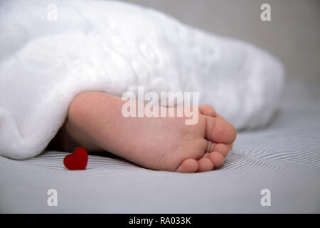 The baby sleeps under a blanket, feet out with a small red heart for the holiday Stock Photo