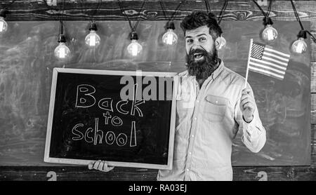 American school concept. Teacher holds blackboard with written phrase back to school and flag of USA. Man with beard on smiling face welcomes to american school, chalkboard on background. Stock Photo