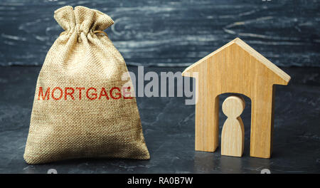 A bag with money and the word Mortgage and a wooden house with a tenant. The accumulation of money to pay interest rates on mortgages. Buying a proper Stock Photo