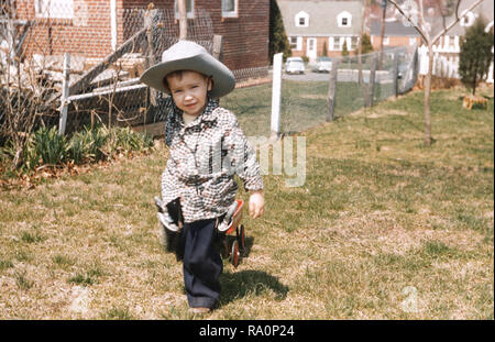 1950s four year old boy is dressed up in his cowboy outfit pulling his wagon. USA Stock Photo