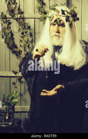 Bearded senior man wizard in long white wig vine crown as Zeus god with medallion on chain indoor on wooden background Stock Photo