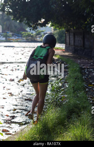 Smiling pretty girl in a sporty dress and backpack holding a coconut with straw with helmet, motorbike and waterfall on background. Puerto Galera, Mindoro Island, popular tourist place in Philippines Stock Photo