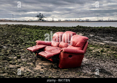 A Red Sofa Dumped on a Rocky Beach with the Heavy Industrial Landscape of Teesside as a Backdrop,  South Gare, Redcar, Yorkshire, England, UK Stock Photo