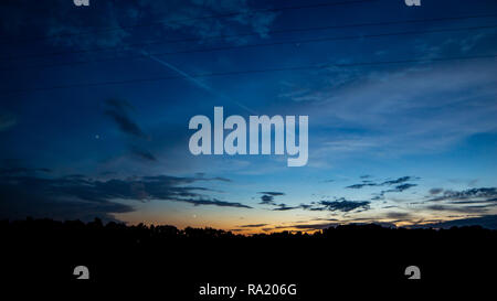 The sun sets behind the tree line while dark islands of clouds float by. Stock Photo