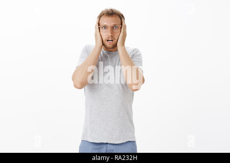 What have I done. Portrait of stunned and shocked intense adult male in glasses holding hands on head gasping and opening mouth from shock making terrible mistake starting panic over white wall Stock Photo