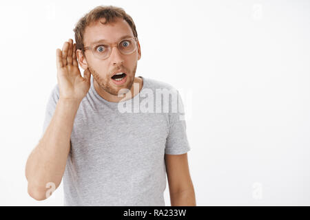 You say what. Intense focused intrigued man with bristle in glasses bending towards with hand near ear and concerned curious look opening mouth from interest listening rumor or gossip over white wall Stock Photo
