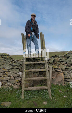 Man in a cap on a hike climbs a stile to cross from one pasture to another in the Shetland Islands of Scotland Stock Photo