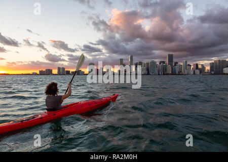 Adventurous girl kayaking in front of a modern Downtown Cityscape during a dramatic sunset. Taken in Miami, Florida, United States of America. Stock Photo