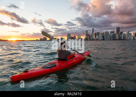 Adventurous girl kayaking in front of a modern Downtown Cityscape during a dramatic sunset. Taken in Miami, Florida, United States of America.