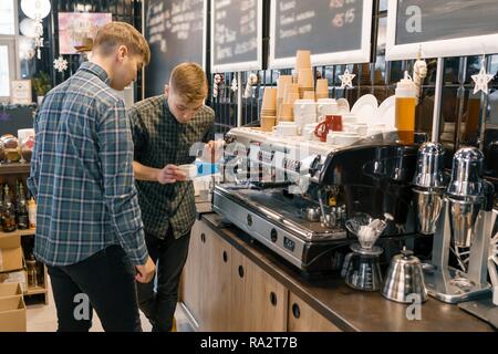 Coffee house, small business, male barista near the coffee machine. Experienced barista coaching teaching young man to brew coffee in professional cof Stock Photo