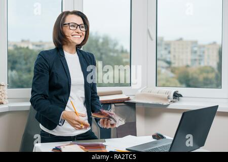 Female decorator interior designer at workplace with fabric samples, laptop. Smiling female in jacket, glasses looking to the side while standing near Stock Photo