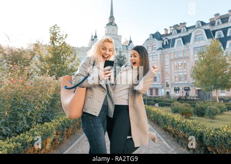 Two young women having fun, looking at the smartphone laughing, sunny autumn day, city background, golden hour. Stock Photo