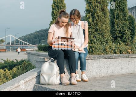 Outdoor portrait of two young beautiful girls students with backpacks, books. Girls talking, looking into a book, urban background. Stock Photo