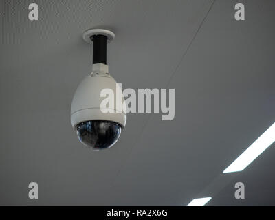 White circular surveillance camera on ceiling in a public setting attached to white ceiling Stock Photo