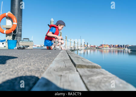 Boy sits on dock fishing wearing red life-jacket looking over edge into Tauranga harbour in marina New Zealand. Stock Photo