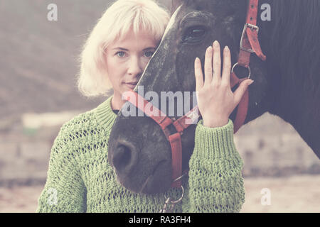 blonde beautiful caucasian model woman with black amazing horse stay together in friendship in fashion style picture concept outdoor nature - hug with Stock Photo