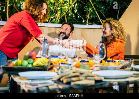 Three caucasian friends having dinner at home by night in the outdoor terrace enjoying food and friendship together having fun - smiles and people eat Stock Photo