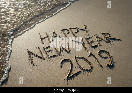 Happy New Year 2019 message handwritten in raised textured letters on a smooth sand beach with oncoming wave Stock Photo