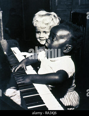 Two children are sitting at a piano. The African American child is playing the piano and singing as the other child looks on ca. 1950s Stock Photo