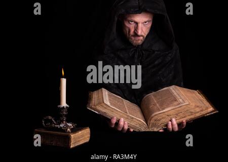 Man, with a black hooded coat, holding an old book in his hands, in front a burning candle, Germany Stock Photo