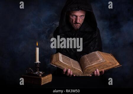 Man, with a black hooded coat, holding an old book in his hands, in front a burning candle, Germany Stock Photo