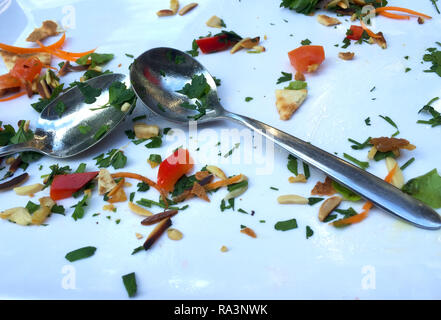 The remians of a meal are on a white plate. This includes tomato, pinenuts, carrot, slivered almonds, parsley and bread. Two silver spoons are on teh  Stock Photo