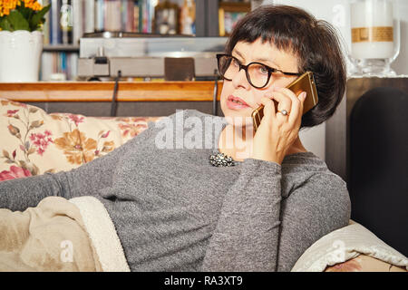 pensive attractive elderly woman lying on the couch and talking on the phone at home. Stock Photo