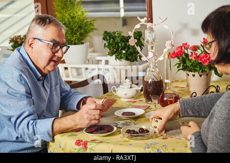 Happy joyful senior couple playing cards and enjoying a snacks at home. Stock Photo
