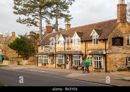 Man and woman walking past the Horse and Hound pub / inn in the picturesque Cotswold village of Broadway with its Cotswold stone buildings and houses Stock Photo