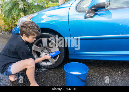Boy earning pocket money cleaning blue compact car with hose, bucket of water and car brush cleaning wheels. Stock Photo