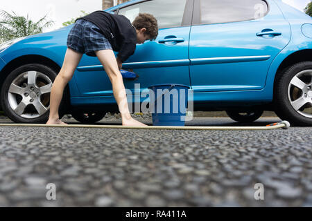 Boy earning pocket money cleaning blue compact car with hose, bucket of water and car brush from low point of view. Stock Photo