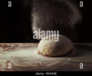 The dough in the shape of a bowl lies on a wooden baking table on a dark black background. Sprinkled with flour. Side view, horizontal photo Stock Photo