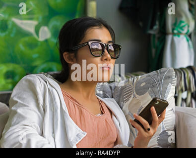 Young Chinese woman holding mobile phone sitting in cafe and looking out of window with green landscape reflected in her sunglasses Stock Photo