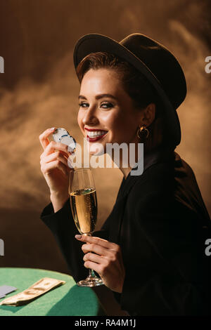 happy attractive girl in jacket and hat holding glass of champagne and poker chips at table in casino, looking at camera Stock Photo