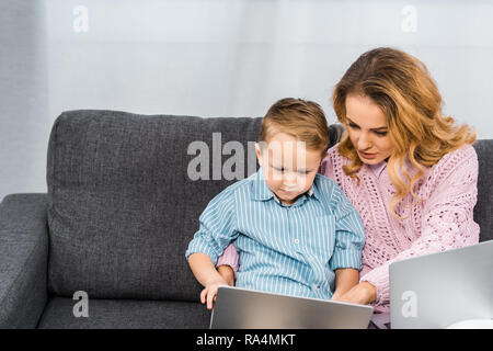 pretty woman with son sitting on sofa and pointing with finger on laptop screen in apartment Stock Photo