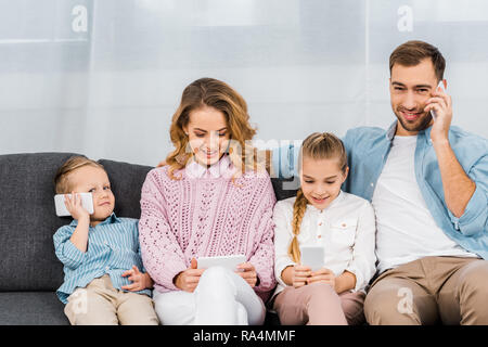 smiling father sitting on sofa and talking on phone while mother and children using smartphones in apartment Stock Photo