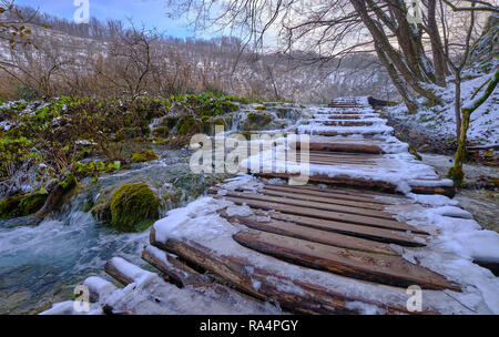 Wooden walking stairs path covered in snow and ice by side of green colored creek. Scene in Plitvice National Park Stock Photo