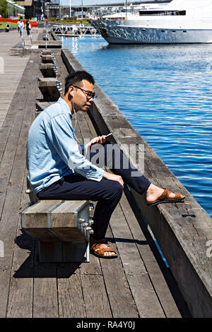 A young Asian man listens to music on his mobile phone in Victoria Harbour in Melbourne Docklands,Victoria Australia. Stock Photo