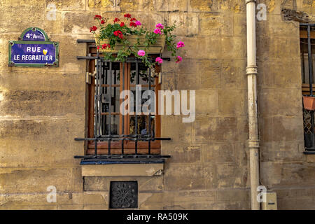A wooden window with metal bars and a flower box on top and a blue street sign on a stone wall in Rue le Regrattier , Ile Saint-Louis ,Paris Stock Photo