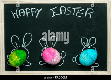 three colorful, pastel Easter eggs with painted rabbit ears against the background of a blackboard Stock Photo
