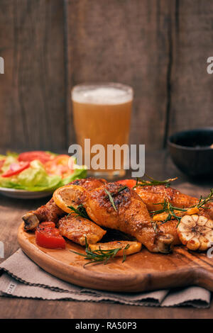 Grilled chicken legs roasted on the grill on wooden chopping board with tomato sauce in a bowl, fresh tomatoes and lettuce leaves, bitter pepper, glas Stock Photo