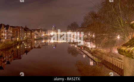 A view of the River Ouse in York just before dawn.  The Ouse Bridge is in the distance with its lights reflected in the still water.  The buildings an Stock Photo
