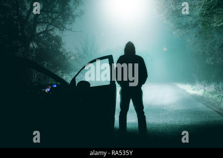 A lone, hooded figure standing next to a car looking at a light beam coming down from a UFO on a misty,c ountry road silhouetted at night by car headl Stock Photo