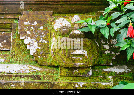 Traditional Bali stone sculpture wall decoration covered with moss, lichens and vegetation with red blossoms Stock Photo