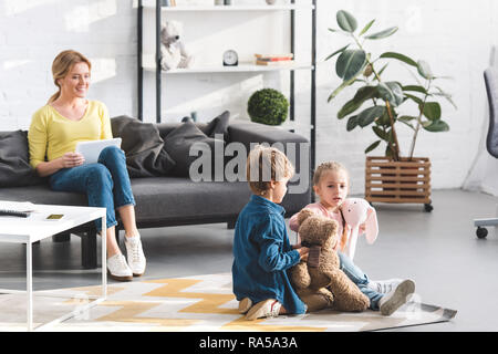 happy mother using digital tablet and looking at adorable children playing with toys Stock Photo