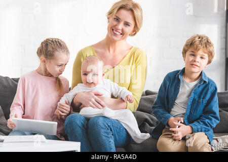 beautiful happy mother smiling at camera while sitting on couch with three adorable kids Stock Photo