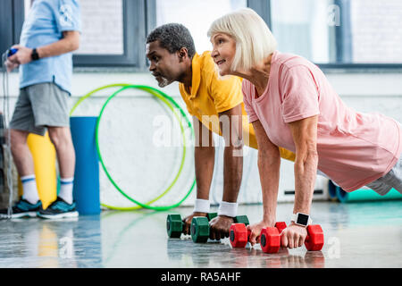 side view of interracial mature couple doing plank with dumbbells at gym Stock Photo